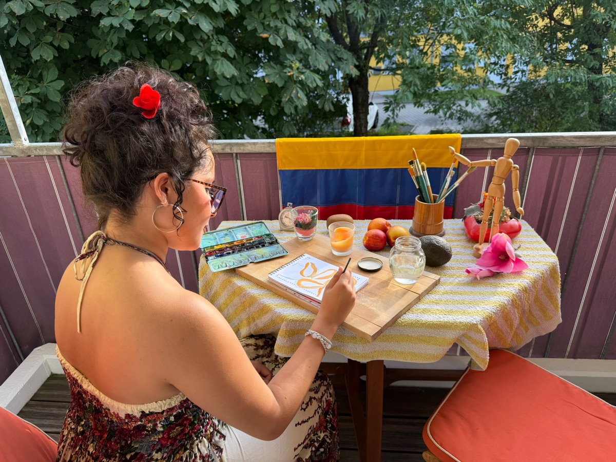 Künstlerin Kim malt die Motiv-Vorlagen mit Aquarell auf ihrem Balkon — mit kolumbianischer Flagge im Hintergrund