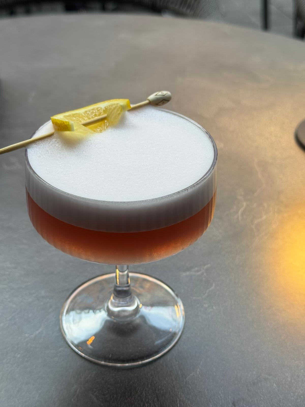 Ein Sour-Cocktail im Coupé-Glas mit Schaumkrone und Zitronenzeste, im warmen Nachmittagslicht beim Hoffest