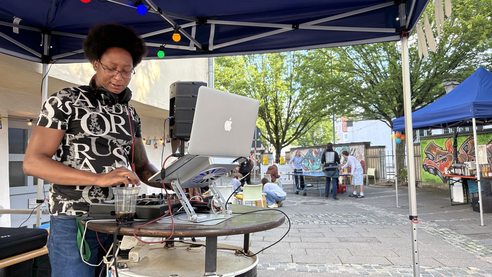 DJ Kenny legt bei WirGestalten im Hof auf, unter blauen Pavillons zwischen Bäumen und Graffiti-Wand